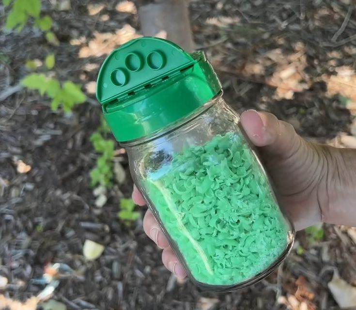 a hand holding a jar filled with green sprinkles on top of dirt