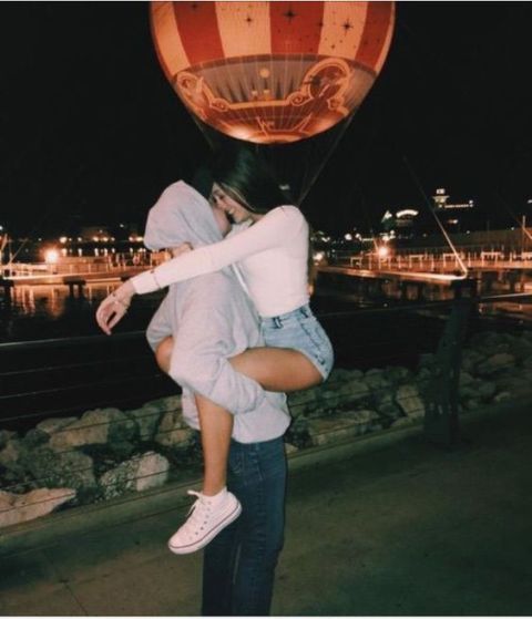 a man and woman hug in front of a hot air balloon that is floating above them