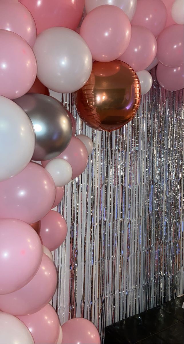 balloons and streamers are hanging from the ceiling in front of a wall that has pink, white and silver confetti on it