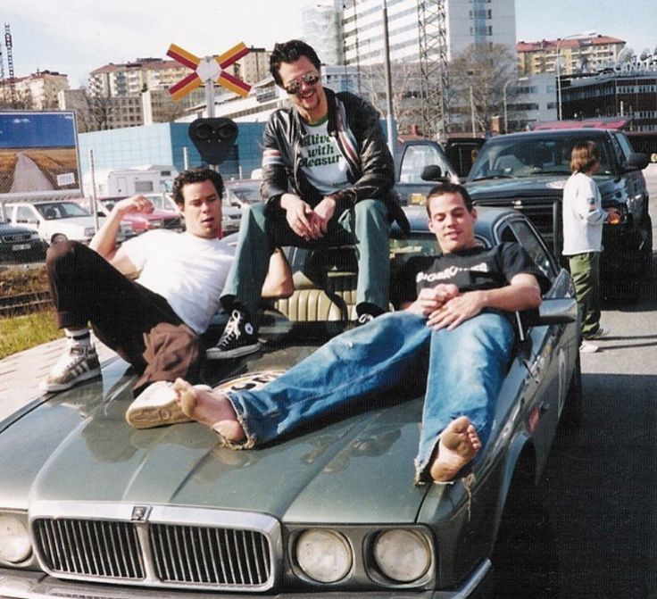 three men sitting on the hood of a car in front of some other people and cars