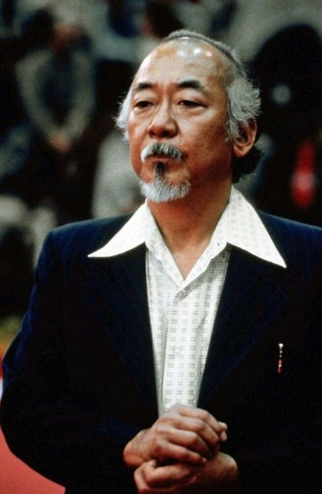 an older man with a mustache standing in front of a crowd wearing a black suit and white shirt