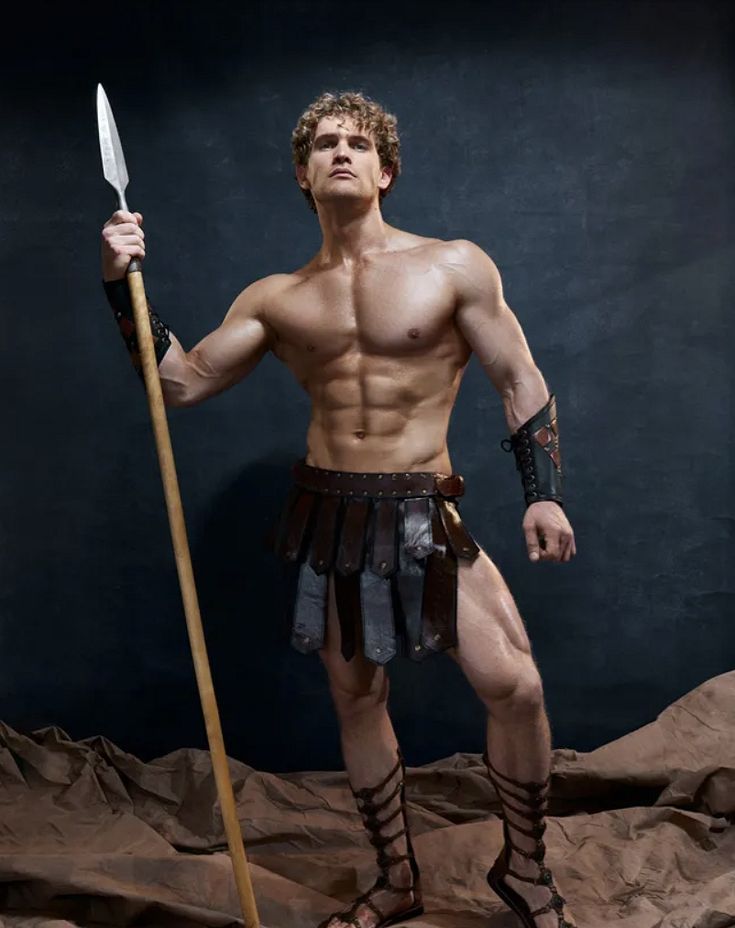 a man with no shirt is holding a spear and posing for a photo in front of a dark background