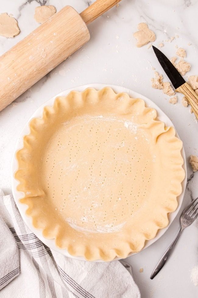 an uncooked pie crust on a white plate next to a rolling pin and utensils