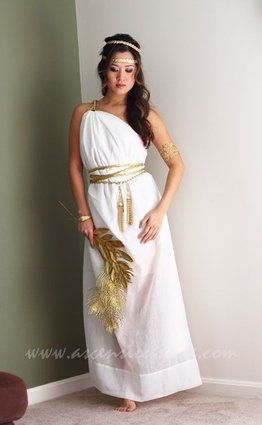 a woman in a white dress with gold accents on her head and arms, standing next to a wall