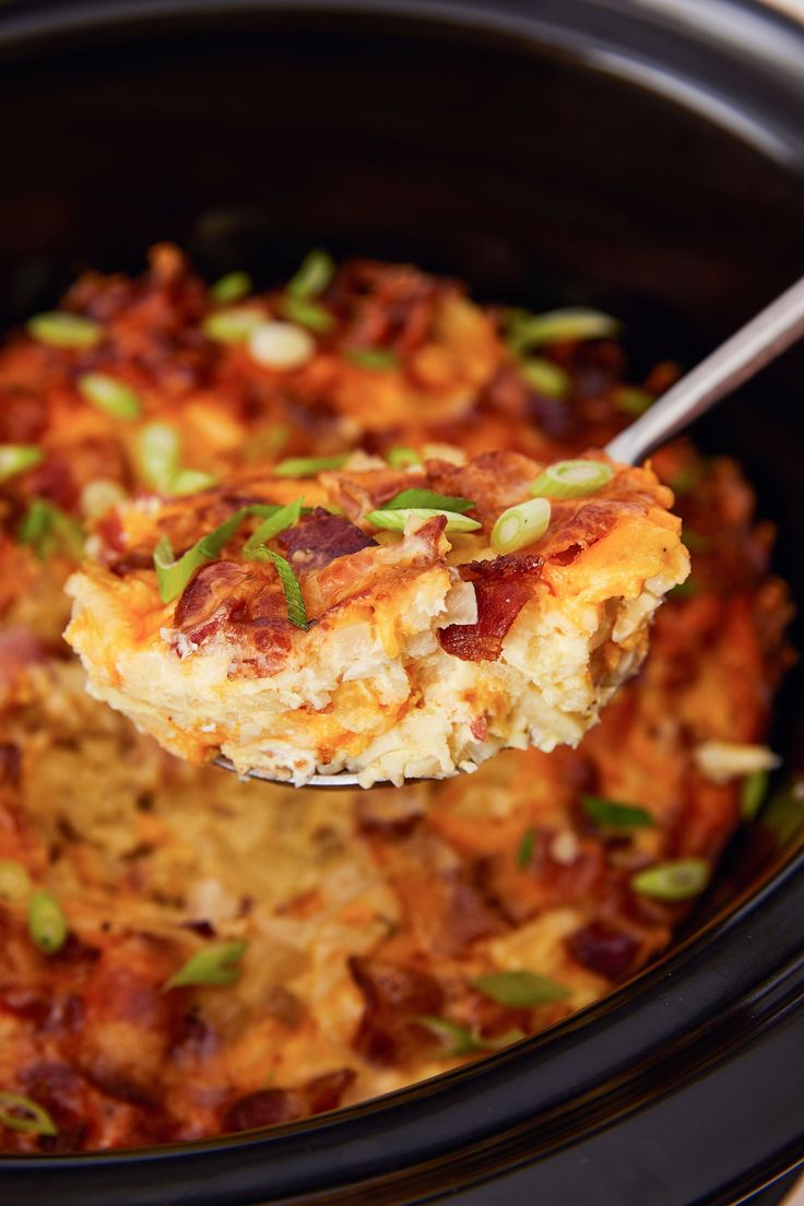 a spoon full of food in a crock pot with green onions and bacon on top