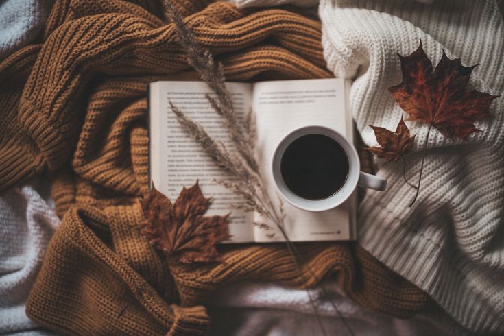 an open book, cup of coffee and autumn leaves on a bed with white blankets