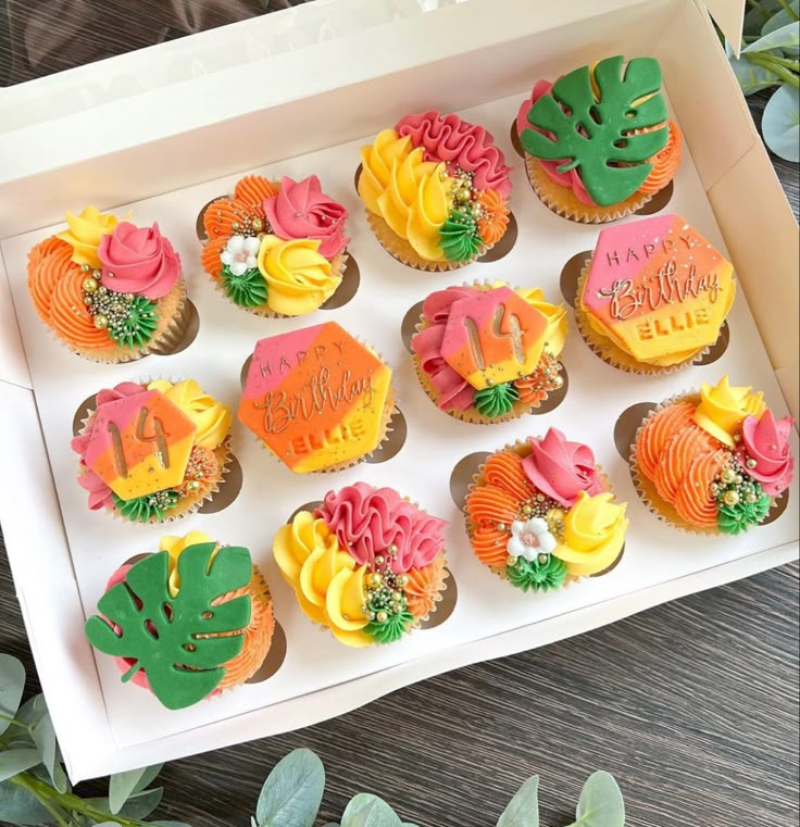 a box filled with cupcakes covered in frosting and decorated with tropical leaves