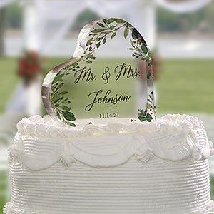 a wedding cake with an acrylic heart on top and greenery in the middle