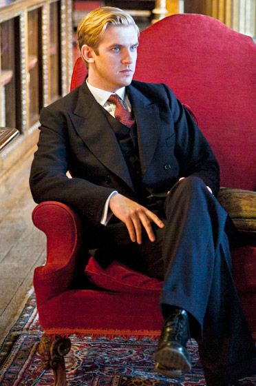 a young man in a suit sitting on a red chair with his legs crossed and looking off to the side