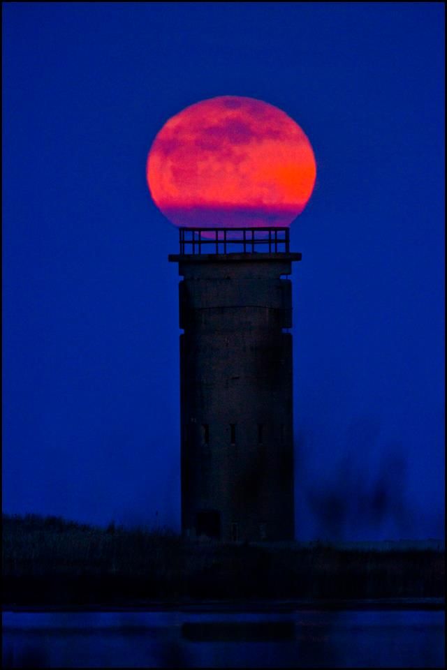 the full moon is setting behind a tower with a red light on it's top