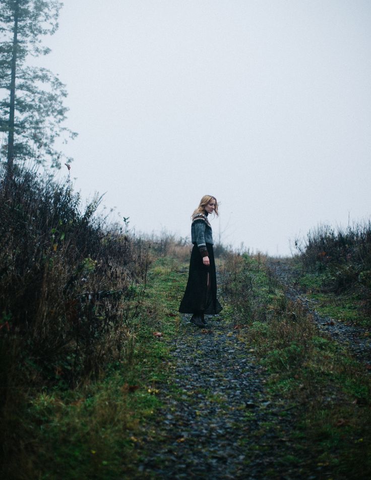 a woman standing in the middle of a field on a foggy day wearing a long black dress