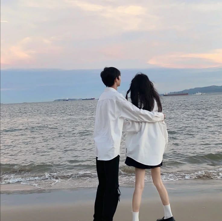 two people are standing on the beach facing the ocean with their arms around each other