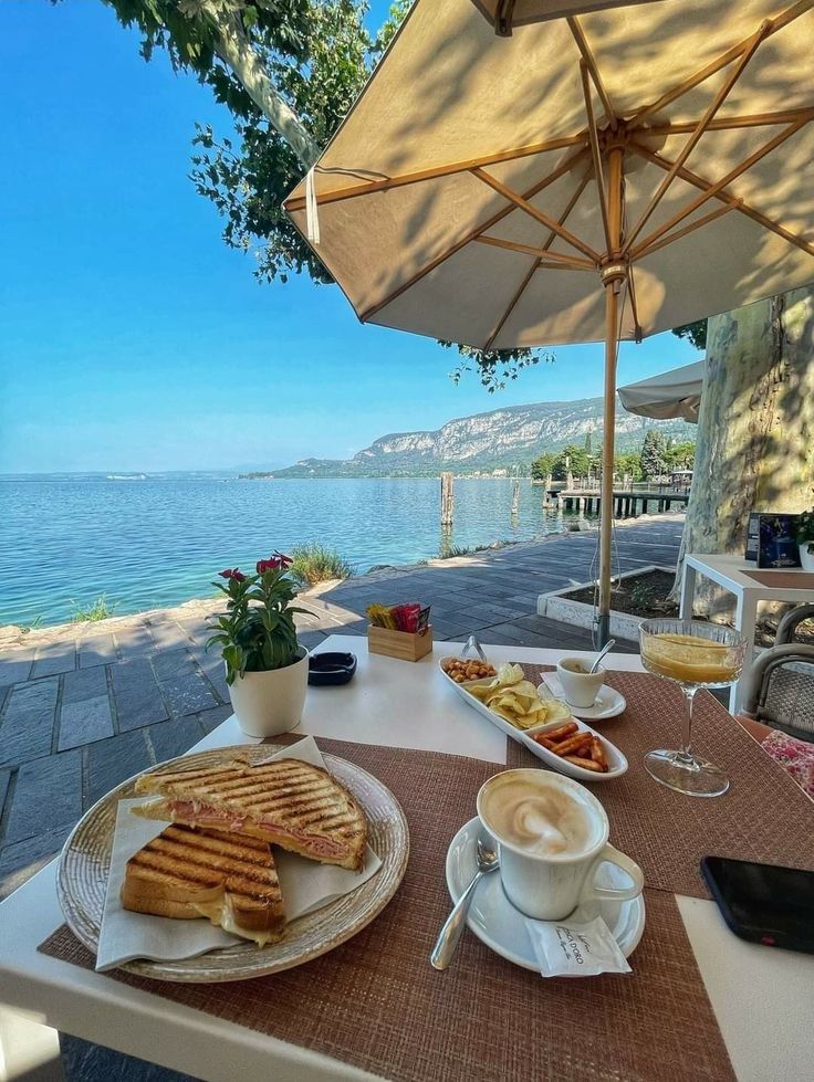 an outdoor table with food and drinks on it next to the ocean, under an umbrella