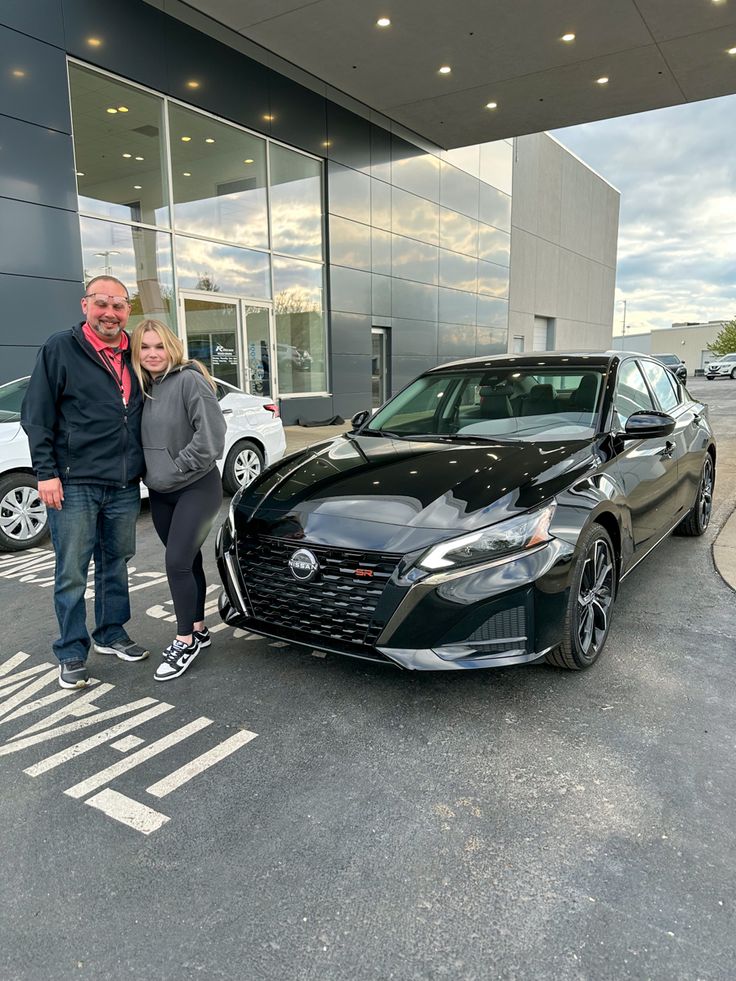 a man and woman standing next to a black car in front of a dealership