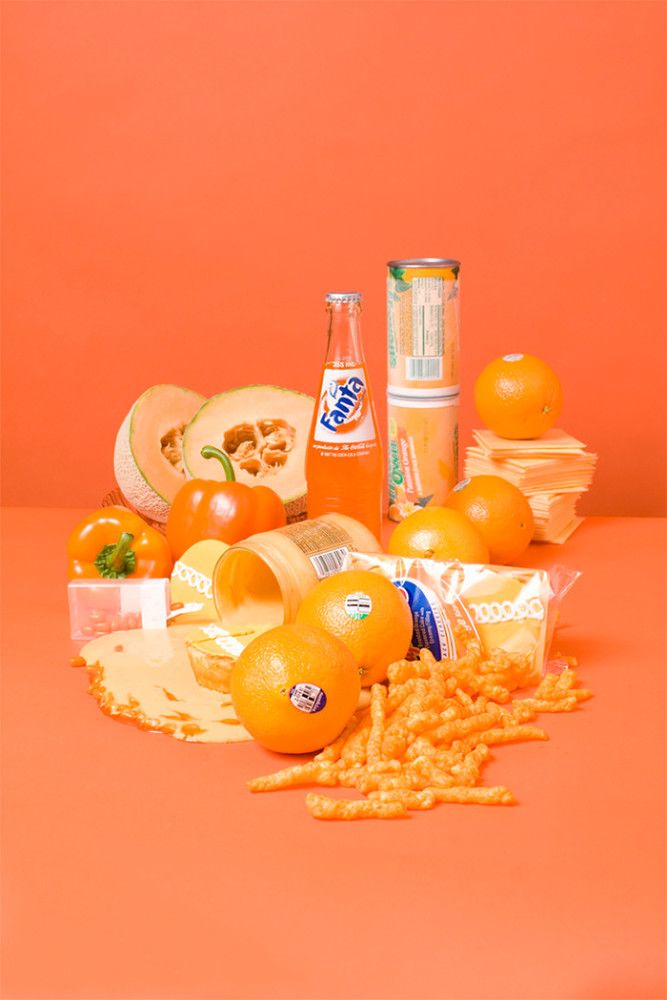 oranges and other food items on an orange colored surface with two cans of soda in the background