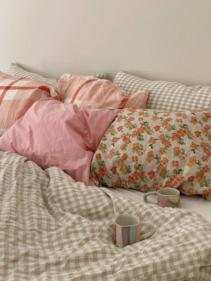 an unmade bed with pink and white pillows, plaid blankets and coffee mugs on it