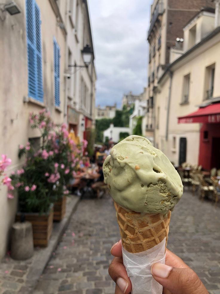 a hand holding an ice cream cone in front of a building on a cobblestone street