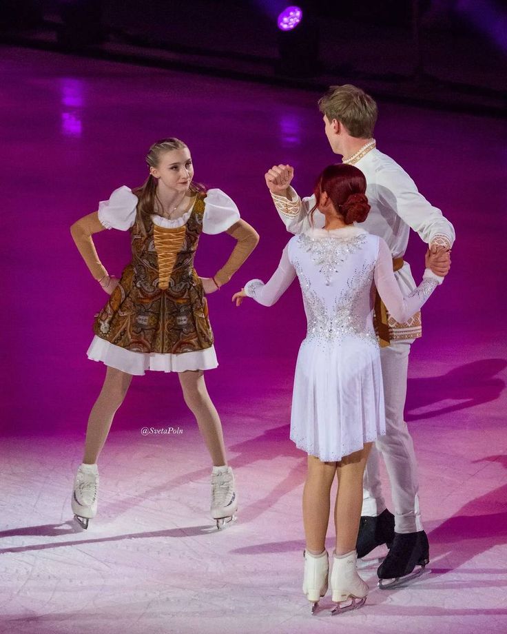 three people are performing on the ice skating rink at an indoor arena, one is wearing a white dress and the other has brown hair