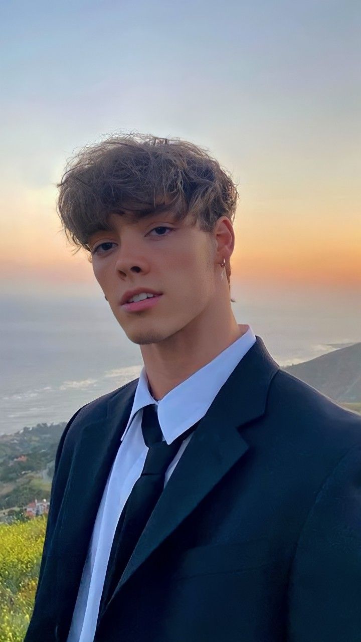 a young man wearing a suit and tie standing in front of the ocean at sunset