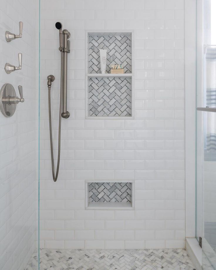 a bathroom with white tile and glass shower doors, two shelves on the wall next to the bathtub