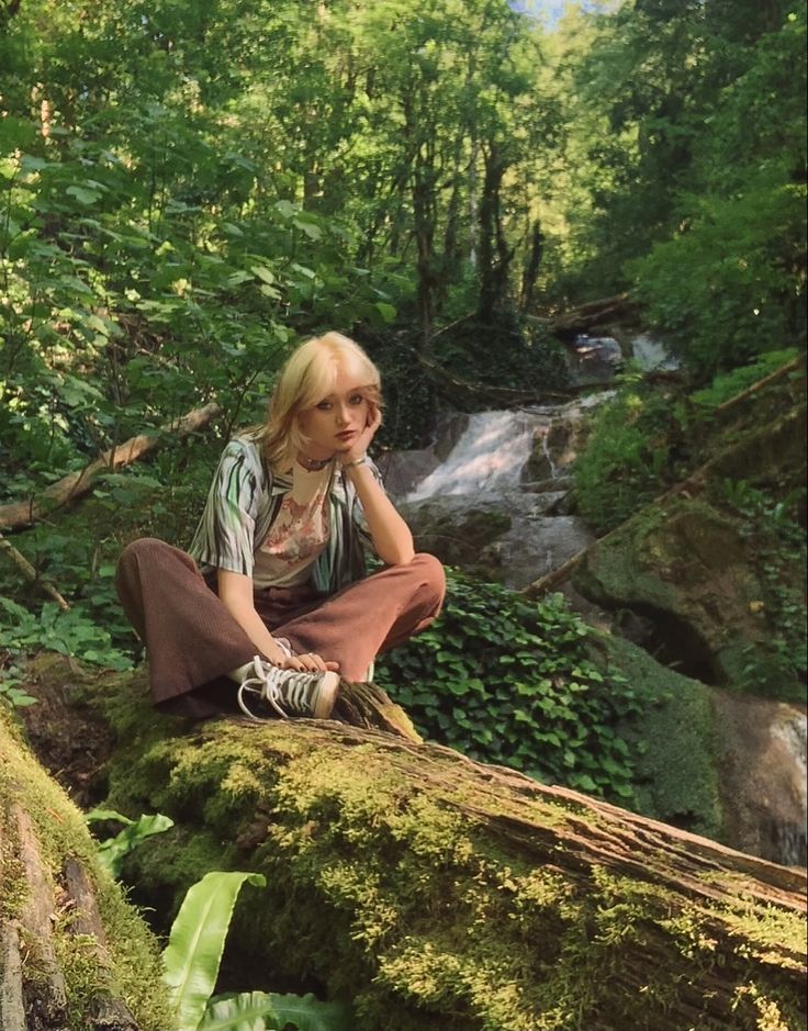 a woman sitting on top of a moss covered rock next to a stream in the forest