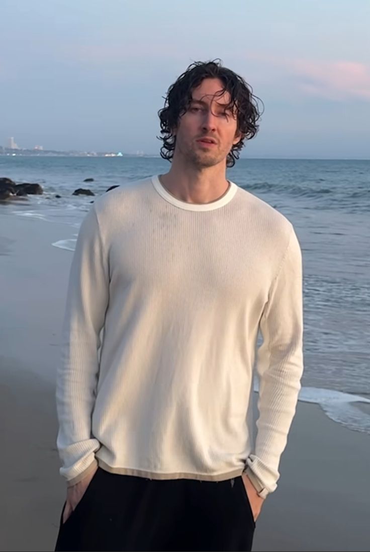 a man standing on top of a sandy beach next to the ocean with his hands in his pockets