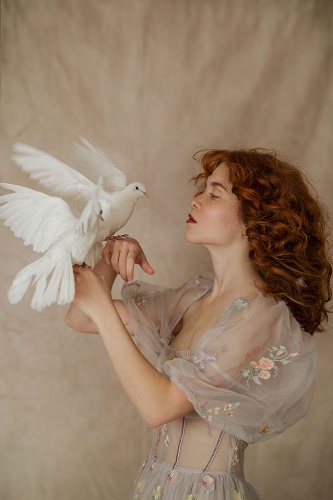 a woman with red hair holding a white dove in her right hand and looking up at it