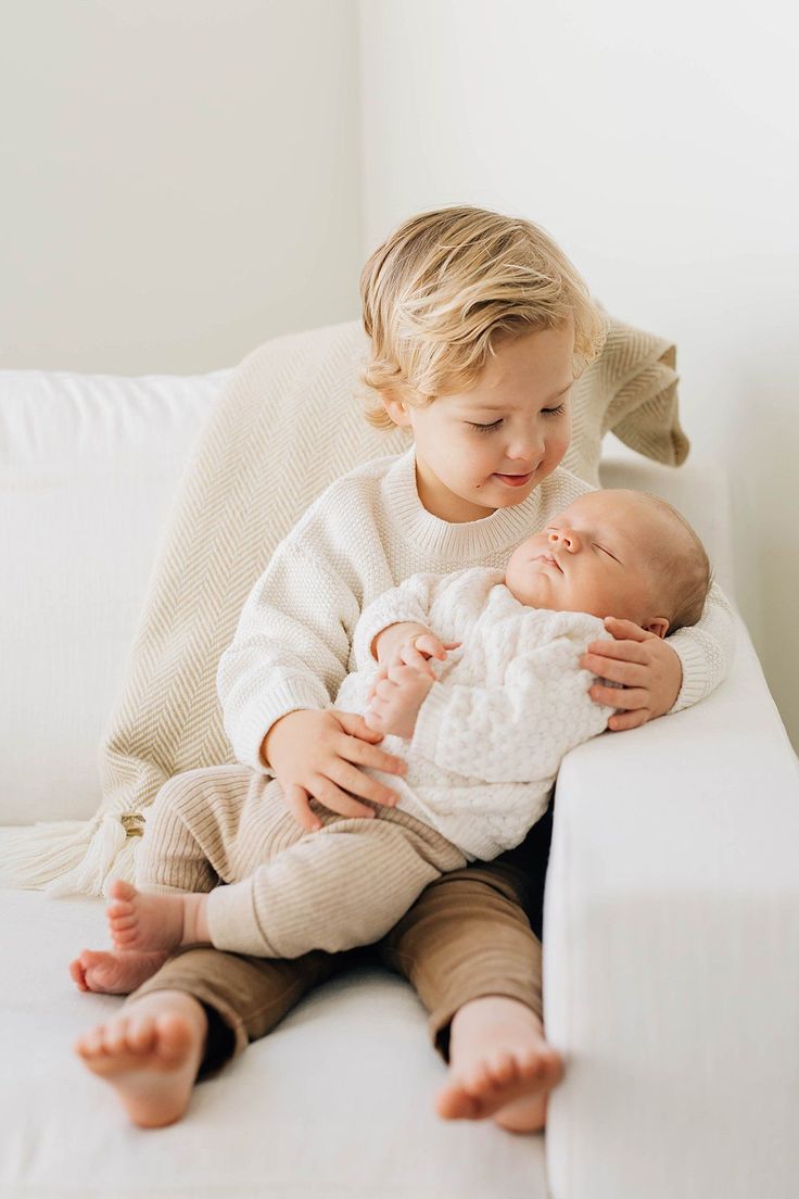 a little boy sitting on top of a white couch holding a baby in his arms