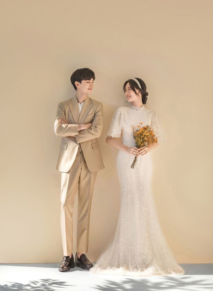 a man and woman standing next to each other in front of a wall holding flowers