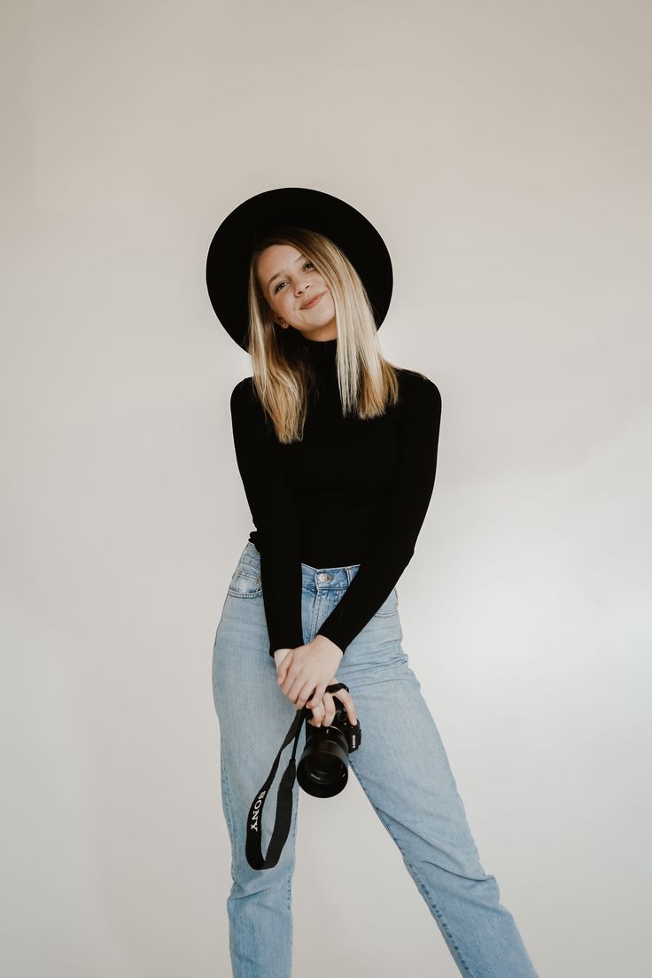 a woman in jeans and a black hat poses for a photo while holding a camera