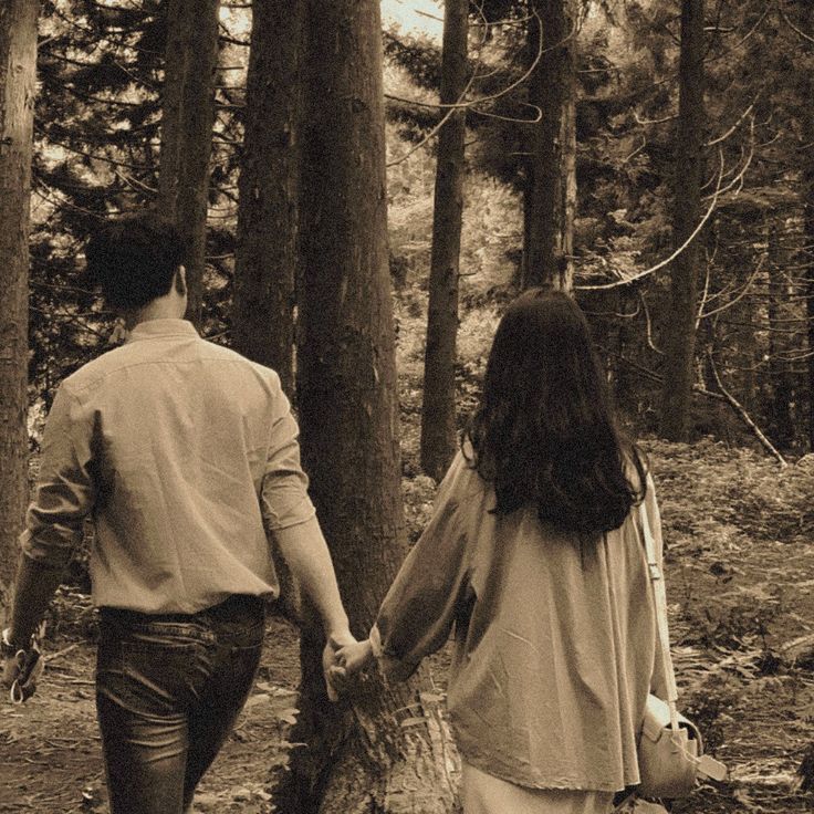 a man and woman holding hands walking through the woods in black and white, with trees behind them