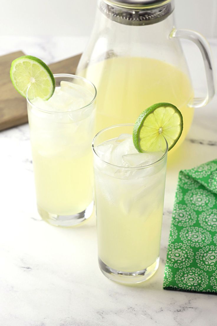 two glasses filled with lemonade and limes next to a pitcher of lemonade