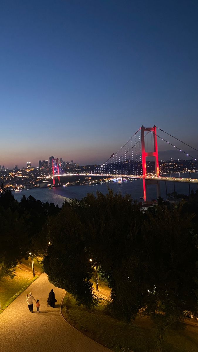 two people are walking down a path near the water at night with bright lights on it