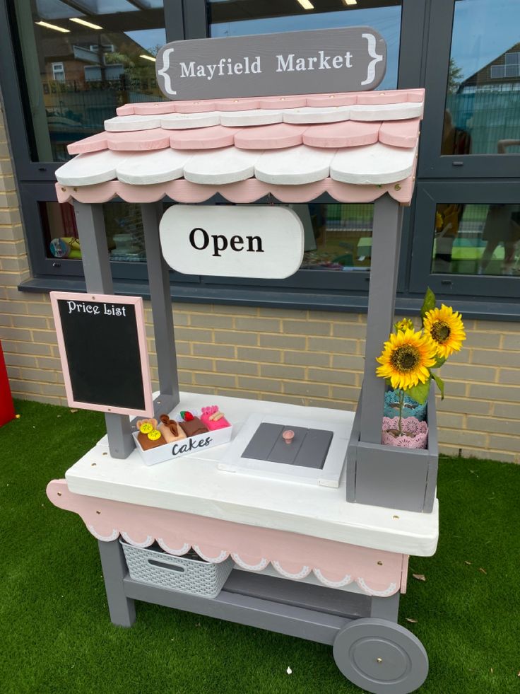 a pink and white cart with flowers in front of a storefront window that says mayfield market open