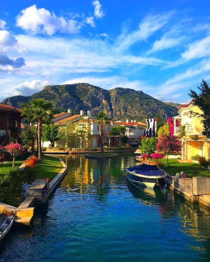 boats are parked on the water in front of some houses and trees with mountains in the background