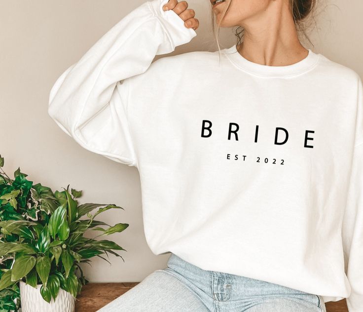 a woman wearing a white sweatshirt with the word bride printed on it, sitting next to a potted plant