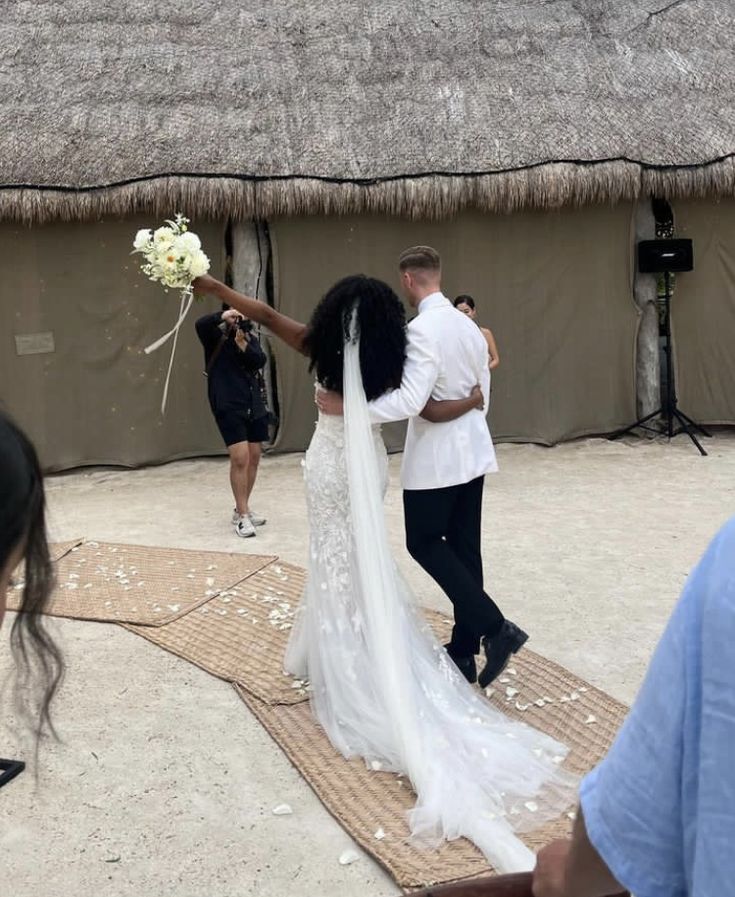 a bride and groom walking down the aisle to their wedding ceremony in front of a thatched hut