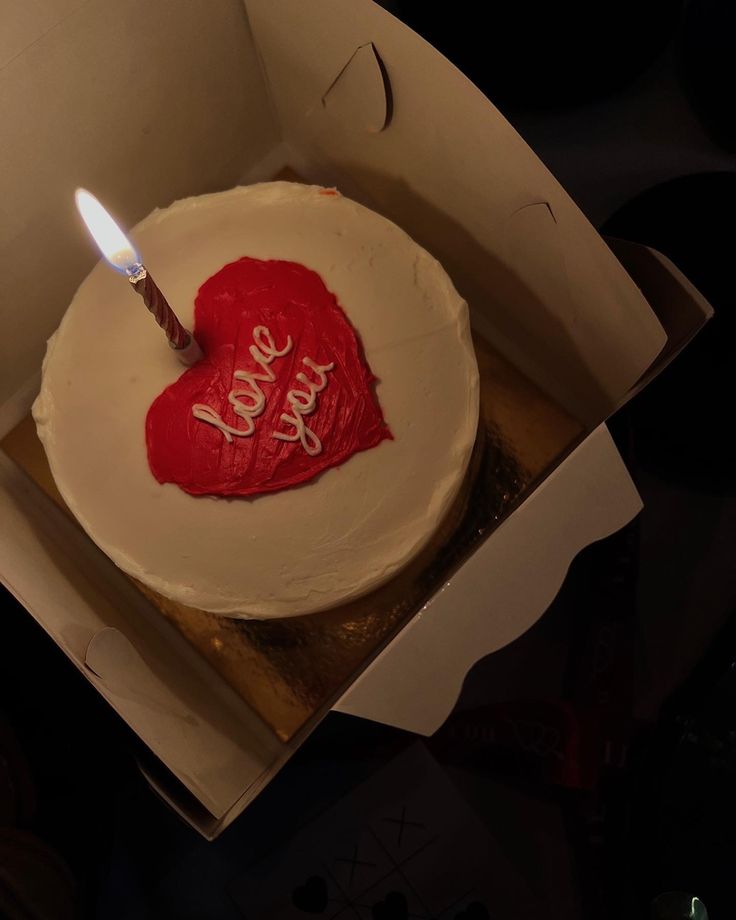 a heart shaped cake in a box with a lit candle on it that says love