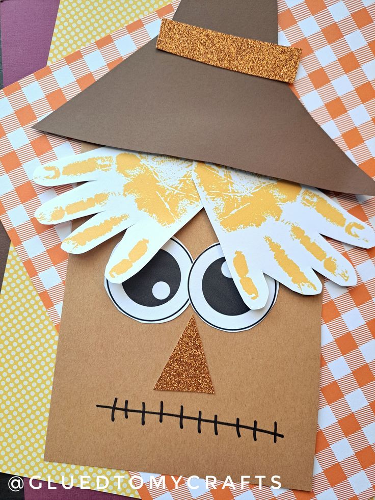 a paper scarecrow craft made with construction paper and glue on top of an orange checkered tablecloth