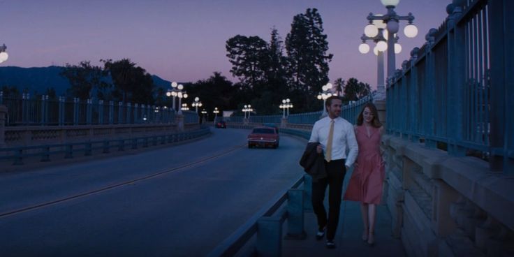 a man and woman are walking down the street at night with their car in front of them