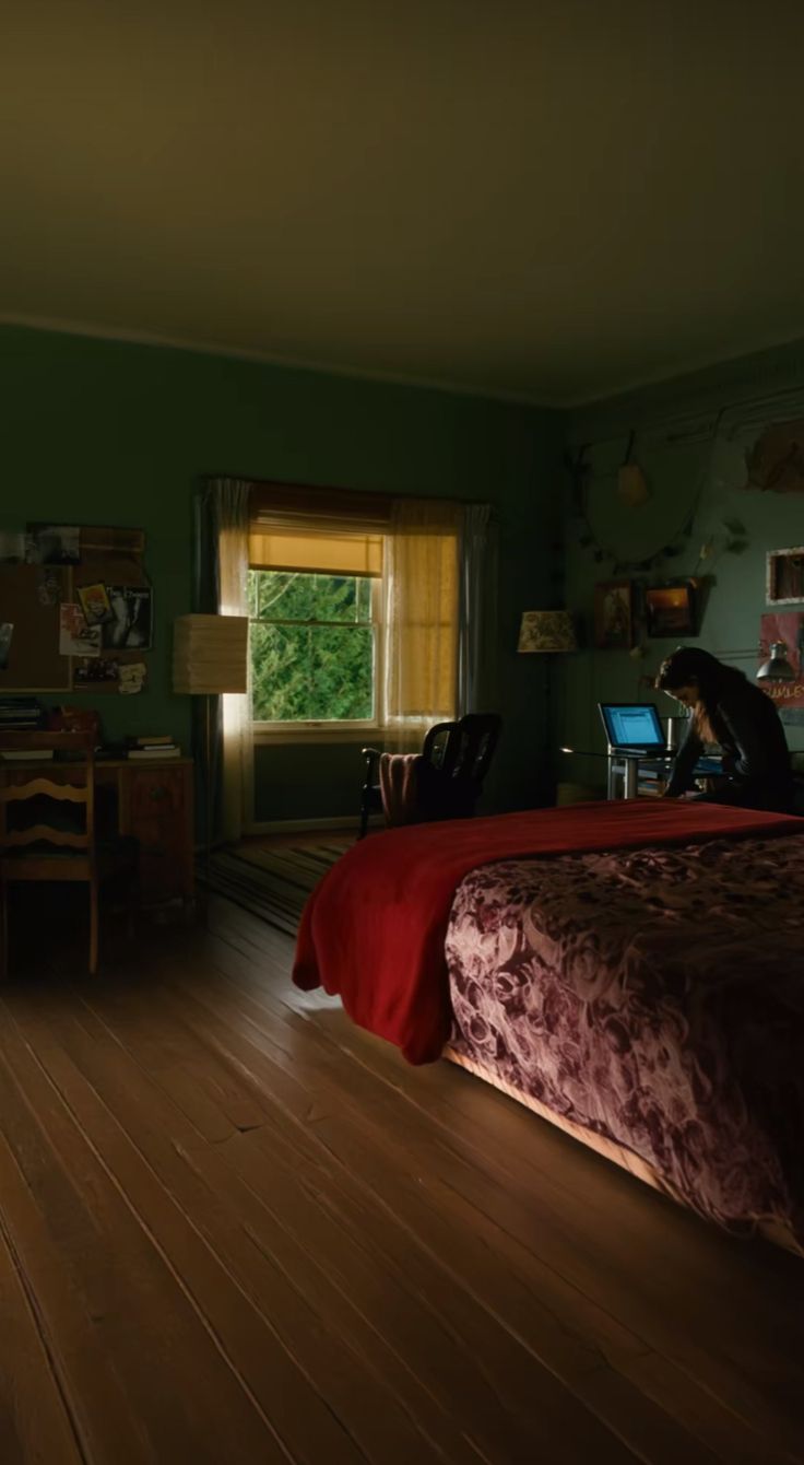 a person sitting on a bed in a room with green walls and wooden floors, using a laptop computer