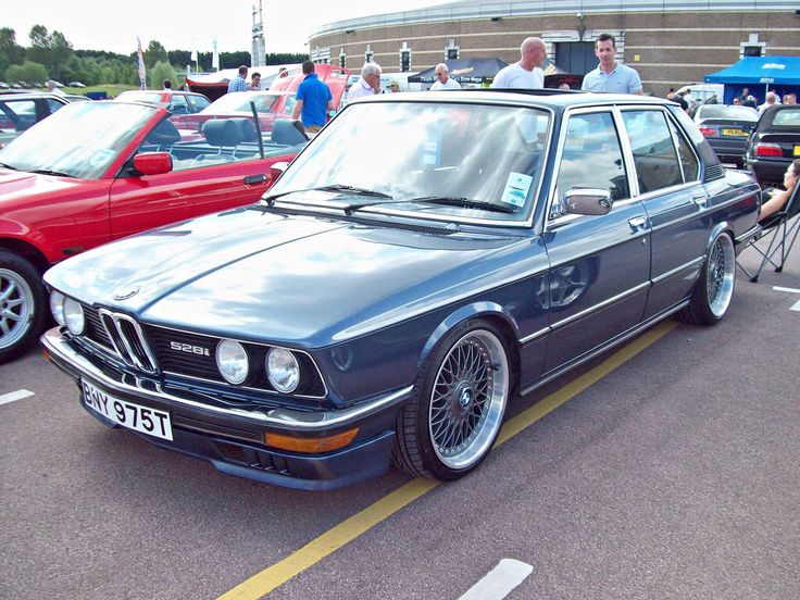 an old bmw is parked in a parking lot with other cars behind it and people standing around