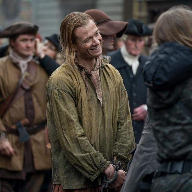 a man with long hair standing in front of other people wearing period clothing and hats