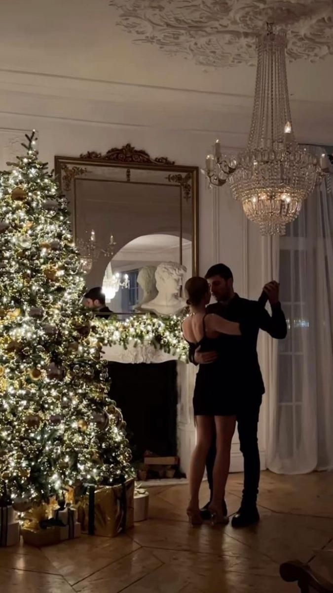 a man and woman standing next to a christmas tree in a room with lights on