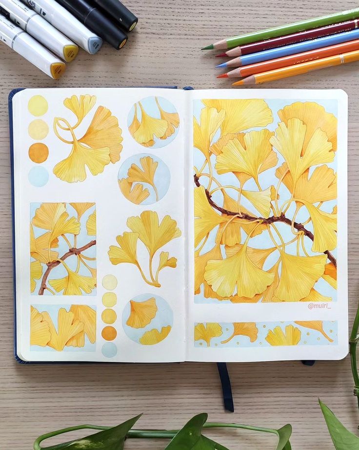 an open notebook with yellow leaves and pencils next to it on top of a table