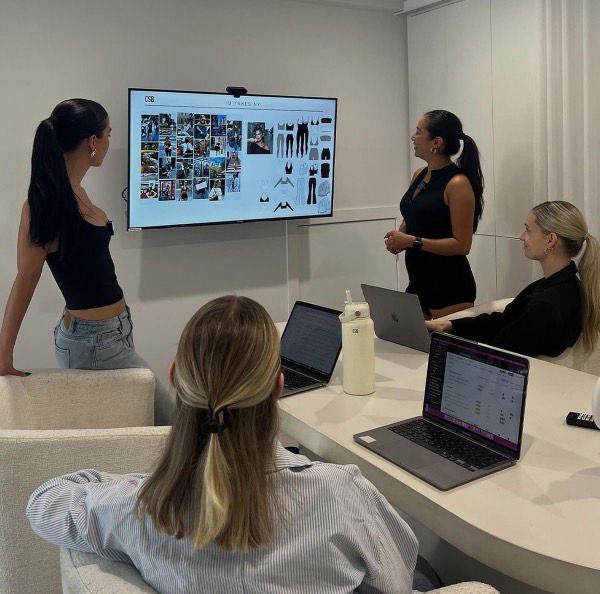 three women sitting at a table with laptops in front of them and one woman standing up