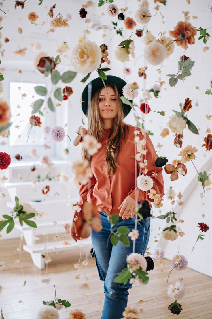 a woman wearing a hat standing in front of flowers and leaves hanging from the ceiling