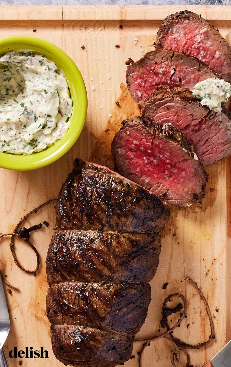 sliced steak on cutting board next to bowl of dip and knife, with small green bowl