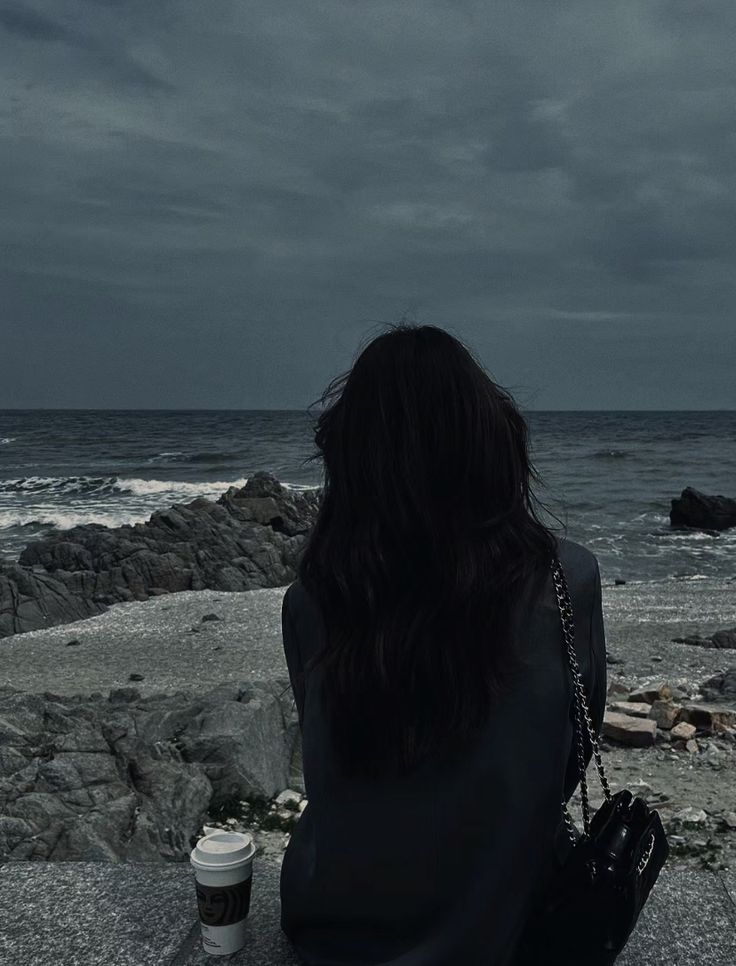 a woman sitting on top of a rock next to the ocean under a cloudy sky