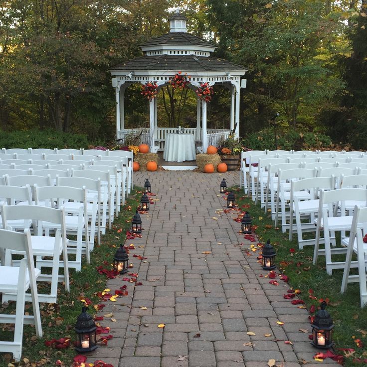 an outdoor wedding setup with white chairs and fall leaves on the ground in front of a gazebo